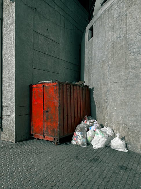 A red metal rubbish container with a weathered, slightly rusted surface, situated against a dark green wall and a concrete wall in an outdoor urban alleyway. The container has vertical ridges and a small lockable door on the front. To its right, on the grey paved ground, there are several loosely tied white plastic rubbish bags filled with waste, with some green and black trash visible through the semi-transparent material. The scene appears to be part of a private waste collection setup, typical of alternative rubbish removal services, with the container positioned for easy access while being tucked away in a narrow alley. Natural daylight casts soft shadows, highlighting the textured surfaces of the container, the bags, and the surrounding walls, emphasizing the functional environment for waste disposal or clearance by an independent rubbish removal company like Waste Disposal Lambeth.