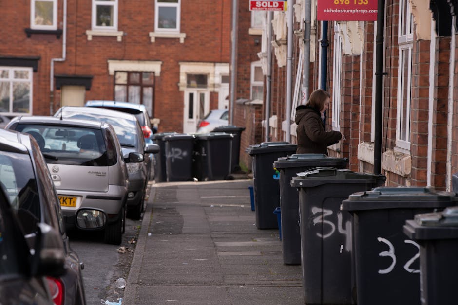 A narrow residential street in an urban area with row houses constructed of red brick, featuring white window frames and stone sills. Several black wheelie bins with lids are lined up along the sidewalk, positioned close to the houses' facades, with some bins displaying white painted numbers or marks. The bins are made of plastic with a matte finish, and their lids are slightly curved. A woman dressed in a dark brown coat and jeans is standing next to one of the bins, facing the wall of the building, appearing to be in the process of managing waste disposal. Parked cars, mostly silver and grey, are aligned along the curb on the left side of the street, with visible side mirrors and rear lights. The environment is illuminated by natural daylight, and the scene appears to be part of a typical urban waste collection area, supporting private rubbish disposal services such as independent collection or on-site clearance, as seen through the lined bins and their arrangement alongside private residences. Waste Disposal Lambeth utilizes such setups for efficient rubbish removal services in the area.