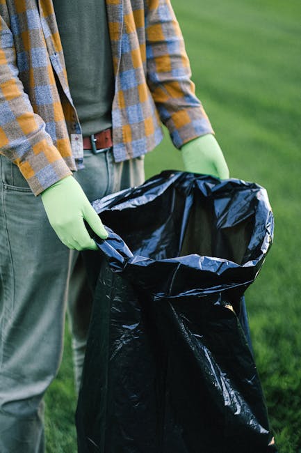 A person standing outdoors on a grassy surface, wearing a blue and yellow checkered shirt over a green top and grey trousers, is holding open a large black rubbish bag lined with a shiny black plastic inner layer. The person is wearing light green gloves, indicating that they are engaged in waste collection or clearance. The background features a well-maintained lawn, suggesting a garden or park environment. The scene emphasizes manual waste handling, which may relate to independent collection or private disposal efforts typical in garden clearance tasks, reflecting the type of service offered by Waste Disposal Lambeth in rubbish removal. The lighting is natural and evenly distributed, with no shadows or artificial elements visible, allowing a clear view of the material and texture of the rubbish bag and the surrounding environment.