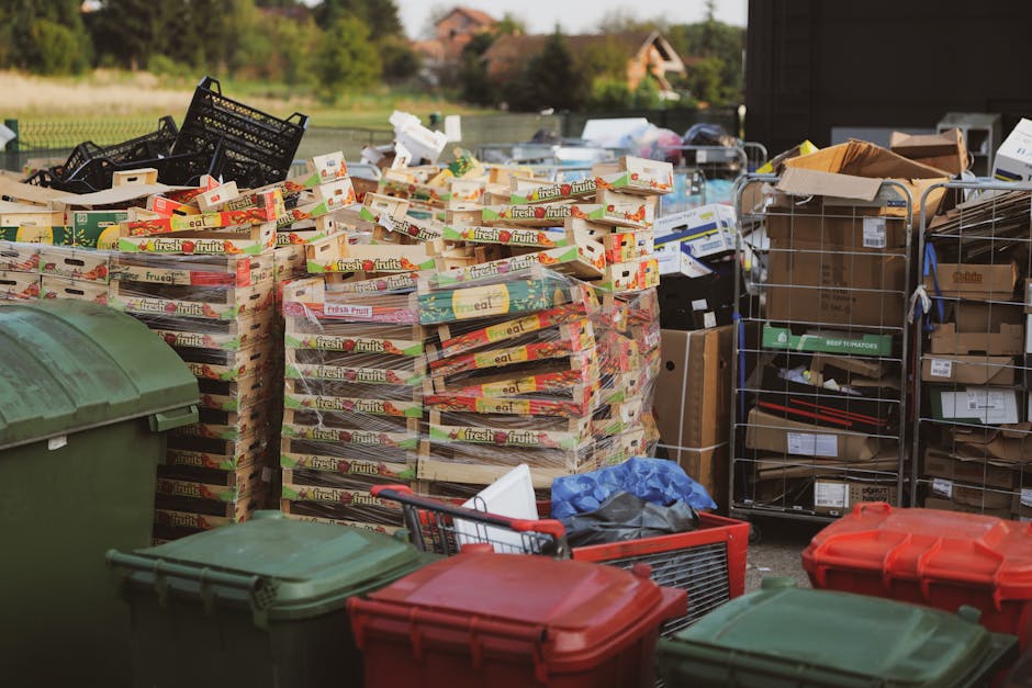 The image displays an outdoor scene with a large stack of empty fruit and vegetable crates, predominantly made of plastic, arranged on a paved surface. The crates are multicoloured, with red, yellow, green, and white tones, featuring the branding 'fresh fruits' prominently on their sides. Behind the crates, there are metal wire storage racks filled with various cardboard boxes and packaging materials, some of which are sealed, and others appear to be partially unpacked or discarded. To the left, a large green plastic wheelie bin with a closed lid is partially visible, with additional similar bins in front, some with red and green lids. Scattered around are a few smaller items, like a blue plastic bag and a red shopping trolley, indicating a collection or clearing process, possibly part of an alternative waste management or private disposal method supported by Waste Disposal Lambeth. The background features a rural or suburban environment with trees and distant houses under a clear sky, illuminated by natural daylight, creating a neutral outdoor atmosphere suitable for rubbish removal or waste handling services.
