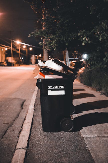 A black wheeled rubbish bin labelled 'ST. JOHN'S' positioned on the edge of a pavement alongside a residential street at night. The bin is partially open and filled with various waste items, including cardboard, paper, and plastic materials. The surrounding environment includes a line of trees with dense foliage casting shadows in the background, and warm street lighting illuminating the scene. The adjacent road is empty, with visible streetlights and some parked cars faintly seen in the distance. This scene reflects a typical private waste collection setup, where refuse is prepared for removal by a waste disposal service, highlighting the importance of timely rubbish presentation to avoid delays in collection schedules. The image emphasizes the practical aspect of on-site rubbish management, often handled independently of municipal schemes, aligning with general principles of private waste handling and disposal services such as those offered by Waste Disposal Lambeth.