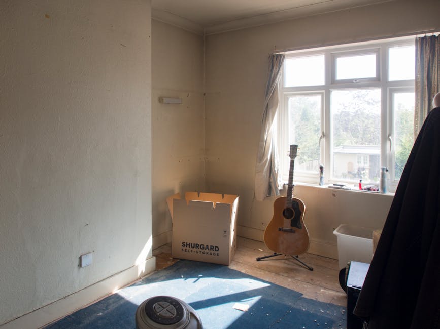 An interior room with beige painted walls and a large multi-pane window allowing natural light to filter in, revealing an outside view of trees and neighboring houses. In the corner near the window, there is a light brown acoustic guitar standing upright on a black stand. Adjacent to the guitar, a cardboard box labeled 'SHURGARD SELF STORAGE' sits on a wooden floor, partially opened. To the right, a white plastic storage bin is positioned on the floor, next to a black garment or coat hanging on a nearby object. A small, circular robot vacuum cleaner rests on a blue carpeted floor in the foreground, with some small debris scattered around. The room appears to be in the process of clearance, consistent with independent or private waste removal, as part of a rubbish clearance service. The environment is bright with natural light, and the scene reflects a typical space prepared for rubbish collection or cleaning, aligned with services offered by Waste Disposal Lambeth.