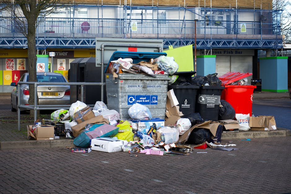 A large pile of mixed waste and rubbish bags, including black plastic bin liners, cardboard boxes, and various discarded packaging, is overflowing from a communal rubbish bin located on a paved street beside a metal railing. The bin is a grey and blue container designated for mixed paper and cardboard waste, with some litter spilling onto the surrounding pavement. Several black, red, and green waste bins are positioned nearby, some partially open with contents visible. Behind the bins, there is a building with storefronts and signage, with one showing a shop front and another with apparent scaffolding or construction work covered in blue netting. A silver car is parked to the left of the waste collection area on the street. The area appears to be in an urban environment with trees lining the sidewalk, and the scene is lit by natural daylight, highlighting the cluttered and untidy state of the waste accumulation. The image reflects a situation where private waste disposal services, such as those provided by Waste Disposal Lambeth, might be involved in rubbish removal from communal or commercial locations.