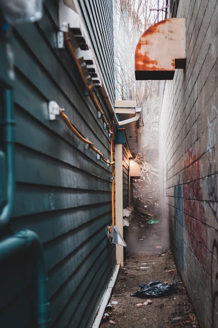 A narrow alleyway between a green corrugated metal building on the left and a grey brick wall on the right, both showing signs of weathering. The alley contains some scattered rubbish, including a black plastic bag and small debris on the dirt ground. Visible outdoor pipes and cables run along the side of the building, with an orange, sun-shaped heat lamp or light fixture mounted on the brick wall near the top right corner. The scene appears foggy or misty, with leafless trees and a muted sky visible at the far end of the alley, suggesting an outdoor, urban environment suitable for private disposal or alternative waste collection arranged by Waste Disposal Lambeth.