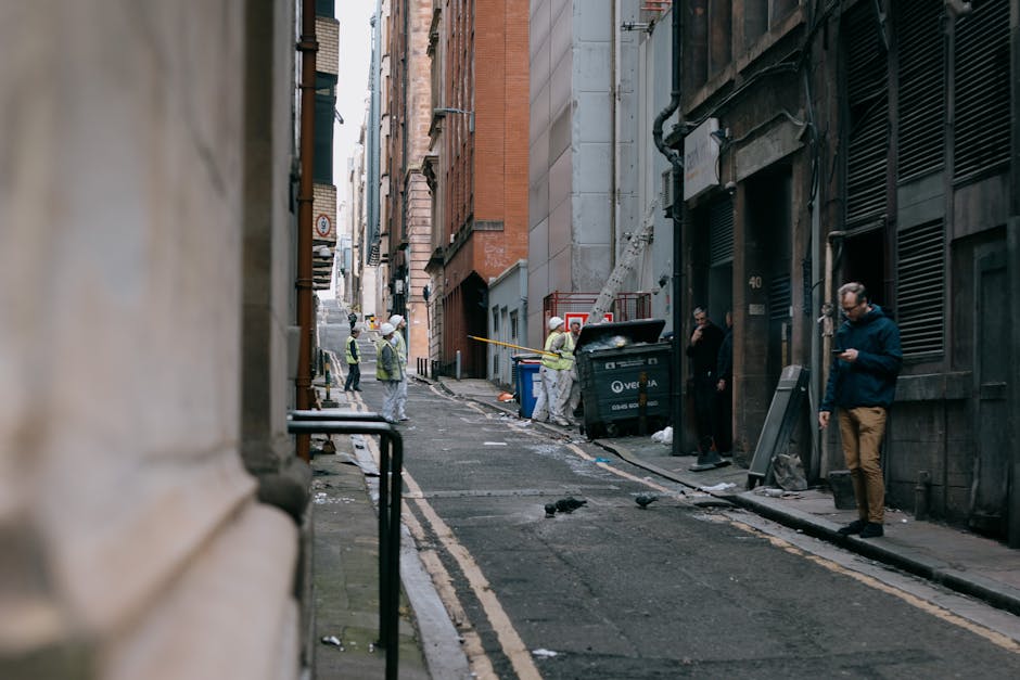 A narrow urban street scene showing a group of workers and individuals near a black waste bin on a steep, cobbled alleyway between tall, multi-storey buildings. The workers, wearing hi-visibility vests and helmets, are engaged in rubbish collection or on-site clearance activities, with some handling debris or equipment. The bin is positioned against a building wall, with clear signs of waste and scattered litter around the area. Adjacent to the bin, a person in dark clothing leans against the building, while another individual in a dark coat stands nearby, looking at their phone. The façades of the buildings display textures of brick and concrete in various shades of brown, grey, and red, with some weathered surface details. The scene is illuminated by natural daylight, creating a neutral, professional observational tone consistent with waste management or private rubbish collection services in a congested, city centre setting, highlighting the challenges of alleyway rubbish access for efficient clearance by Waste Disposal Lambeth.