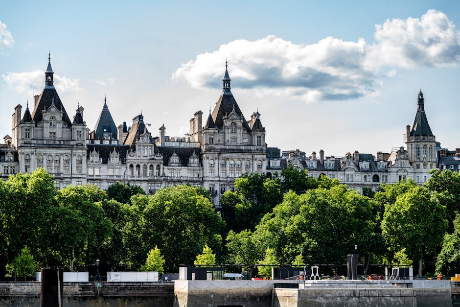 A detailed view of a grand historic building with elaborate architectural features, including towers topped with pointed spires, ornate facades, and numerous windows, set behind a line of lush green trees with dense foliage. The building's stone exterior appears light in colour with a textured finish, displaying intricate decorative elements and a slightly aged appearance. The scene is illuminated by natural daylight, casting gentle shadows and highlighting the structural details of the building against a partly cloudy sky with fluffy white clouds. In the foreground, a river or canal runs along the base of the trees, with dark vertical posts at the water's edge. This setting suggests an urban environment where private waste disposal or on-site clearance services, such as those provided by Waste Disposal Lambeth, can support the management of rubbish generated by events or building maintenance near historic and protected sites, emphasizing the importance of professional rubbish removal in maintaining the area's aesthetic and structural integrity.