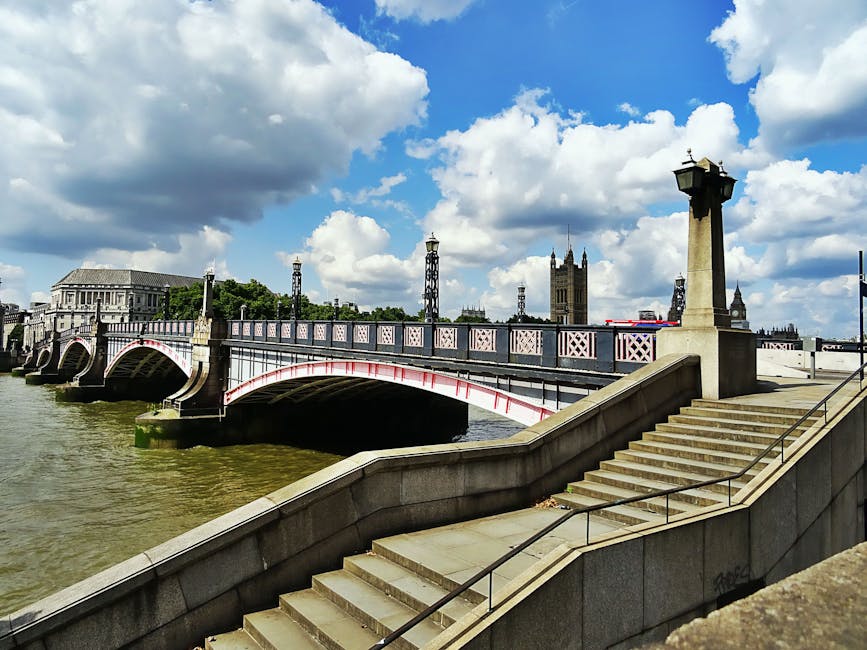 The image depicts a historic bridge crossing a river with a decorative metal railing painted in white and pink accents. The bridge features stone arches supporting its structure, with several ornate lampposts lining the top, casting a subtle shadow on the walkway. On the right side, stone stairs with metal handrails lead down to the riverbank, covered in a smooth, light-colored stone finish. The surrounding environment includes greenery in the background, with trees and official buildings, possibly government or civic structures, visible beyond the bridge. The sky above is partly cloudy with bright white clouds scattered across a vibrant blue backdrop, suggesting daylight and fair weather. The scene is captured with natural lighting, emphasizing the textures of the stone, metal, and water, conveying a calm, urban setting that could be associated with private or alternative waste disposal methods in a city context, indirectly linking to waste management services such as those offered by Waste Disposal Lambeth.