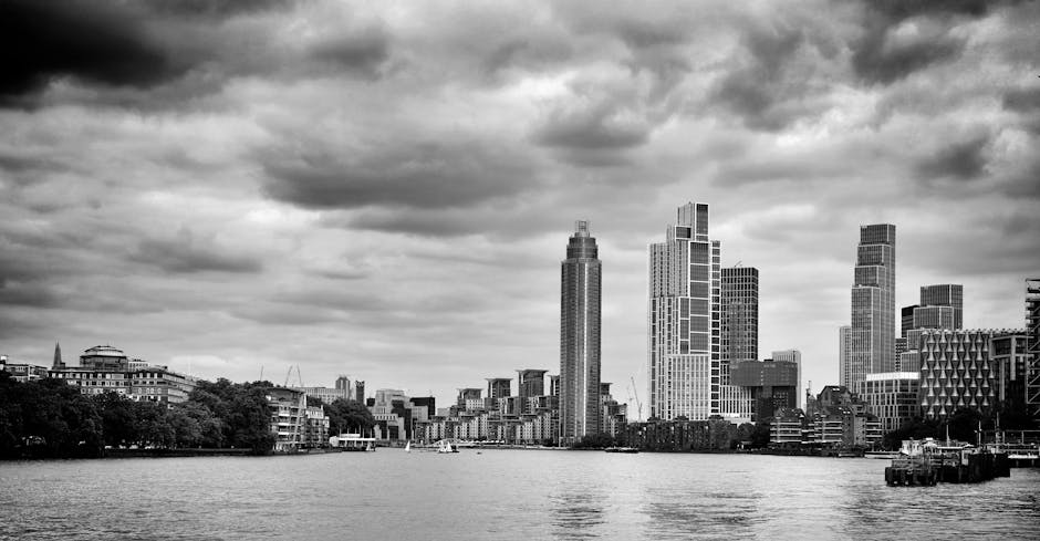 A black and white photograph of a city skyline viewed from across a wide river. Prominent high-rise buildings with varied rectangular and cylindrical shapes dominate the scene, with some featuring distinctive patterns and finishes. The tallest structures are positioned towards the center, while lower buildings line the riverbank on both sides. In the foreground, the calm water of the river reflects the urban landscape, with a boat visible on the water near the right edge. The sky overhead is filled with layered, textured clouds, creating a moody atmosphere. The environment appears urban and developed, with no visible natural or rural elements. The scene relates to urban waste management and the need for efficient rubbish removal services, similar to those offered by Waste Disposal Lambeth, especially when considering the logistical aspects of managing waste in densely populated metropolitan areas.
