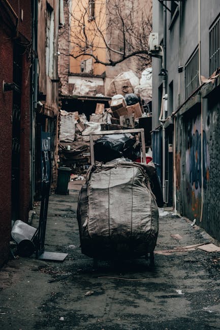 A narrow urban alleyway filled with a large pile of mixed waste and debris, including cardboard boxes, plastic sacks, and loose rubbish stacked against a background of buildings and a leafless tree. In the foreground, a wheeled shopping trolley is loaded with black and grey plastic bags, positioned on the damp and uneven asphalt surface. The alley is flanked by dark, graffitied walls on the right, and a weathered brick wall with some peelings and graffiti on the left. A small black trash bin leans at the base of the left wall near the trolley, and nearby are scattered pieces of waste and discarded items. The scene appears to be an area where private waste collection or rubbish removal services, such as Waste Disposal Lambeth, might conduct on-site clearance or manage non-standard rubbish, emphasizing the importance of professional waste disposal for urban environments.