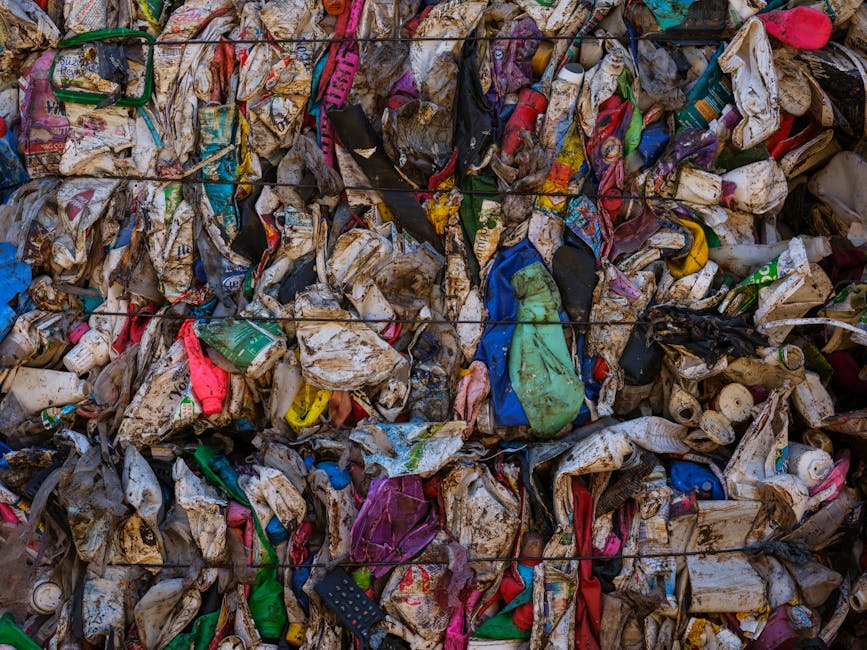 A large pile of compressed household waste and rubbish, consisting mainly of crumpled and torn plastic packaging, paper cartons, and discarded single-use items, appears stained and dirty, with some pieces showing signs of exposure to dirt and moisture. The waste is tightly compacted and stacked outdoors, likely for transportation or disposal, with various colors of plastic and paper materials visible, including green, pink, blue, and black. The texture indicates a mix of soft and rigid materials, with some plastic items still identifiable by their smooth finishes, while paper components are crumpled and stained. The background is not clearly visible, but the waste occupies most of the frame, suggesting an industrial or waste collection site. The scene has a dull lighting, typical of overcast or shaded conditions, emphasizing the grimy and cluttered nature of the rubbish, which is typical of items prepared for private rubbish removal services such as Waste Disposal Lambeth, focusing on bulky waste and refuse collection for recycling or disposal purposes.