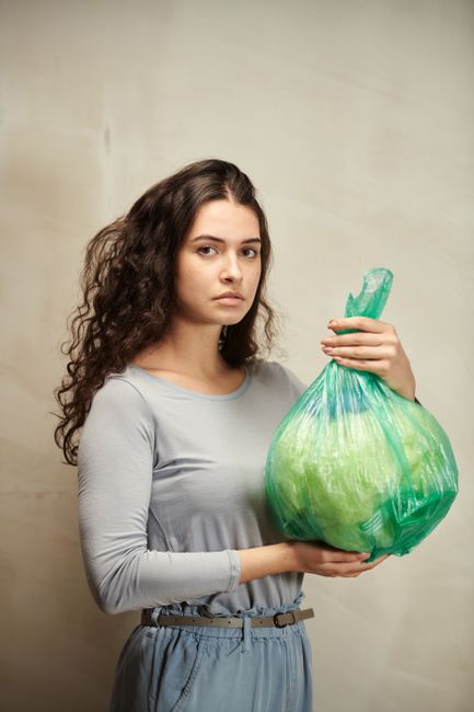 A young woman with long, curly brown hair and light skin is standing indoors against a neutral beige background. She is wearing a light grey, long-sleeved top and high-waisted grey trousers with a thin belt. She has a neutral facial expression and is holding a large, green plastic rubbish bag with both hands, gripping its top gathered section. The bag appears semi-transparent, with a textured surface, and is filled with waste or debris, causing it to be bulging and slightly uneven. The setting suggests an environment related to household waste collection or outdoor rubbish removal, which might be connected to private waste disposal services like Waste Disposal Lambeth. The lighting is soft and even, emphasizing the textures of the plastic bag and the woman's clothing, in a context that aligns with alternative waste handling or on-site rubbish collection activities.