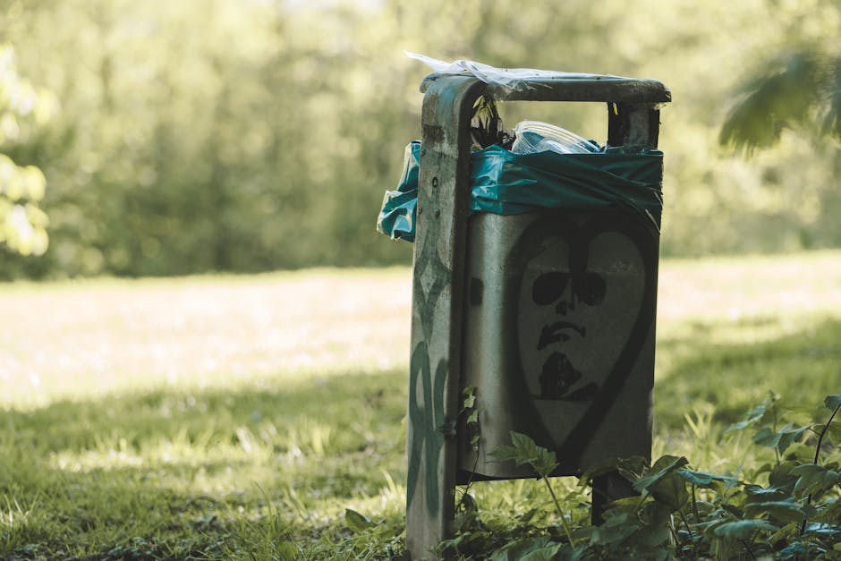 A weathered metal rubbish bin with a graffiti-styled face painted on the front, featuring dark eye sockets, a nose, and an open mouth, situated outdoors on a grassy area. The bin has a black lid, which appears slightly bent or damaged, and is partially covered with clearing bags made of plastic or fabric in shades of blue and green, placed on top. Surrounding the bin are green leaves and a few small plants growing at its base, with a backdrop of blurred trees and sunlight filtering through foliage, creating a natural outdoor setting. The environment suggests an informal or public space, possibly a park or garden, where waste disposal is handled via independent collection methods, aligning with private or alternative rubbish removal services rather than municipal curbside collection. The scene emphasizes the aspect of on-site or site-specific rubbish disposal, reflecting practices of waste management companies like Waste Disposal Lambeth that provide targeted waste removal services outside traditional council systems.