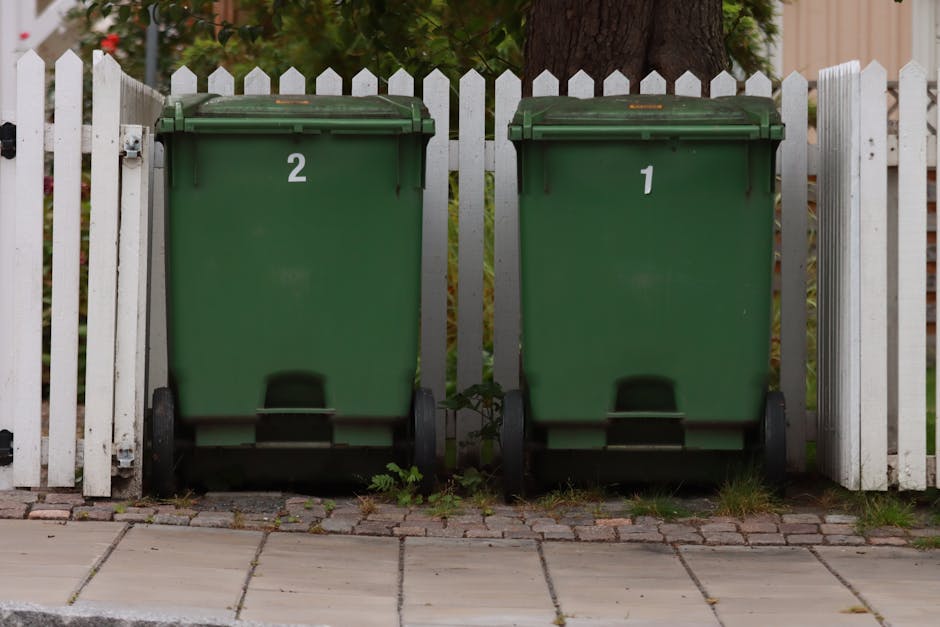 Two green wheeled rubbish bins with black bases, positioned side by side on a cobblestone path in front of a white picket fence, with bin number 2 on the left and bin number 1 on the right. The bins are made of plastic with textured surfaces and slightly domed lids, showing some minor rust or weathering on the edges. Behind the bins, a large mature tree trunk is visible, with green foliage and hints of red flowers in the background. The setting appears to be an outdoor residential area, possibly a driveway or garden boundary, with natural daylight illuminating the scene. The concrete pavement extends across the foreground, with small patches of grass and weeds growing between the stones near the bottoms of the bins, suggesting regular waste collection or the potential for alternative disposal options outside of local council services. The positioning of the bins and the scene's tidy appearance reflect typical private waste management arrangements. Waste Disposal Lambeth specializes in rubbish removal, offering services suited for home or garden clearance, as indicated by the scene's focus on general waste containers in a domestic environment.