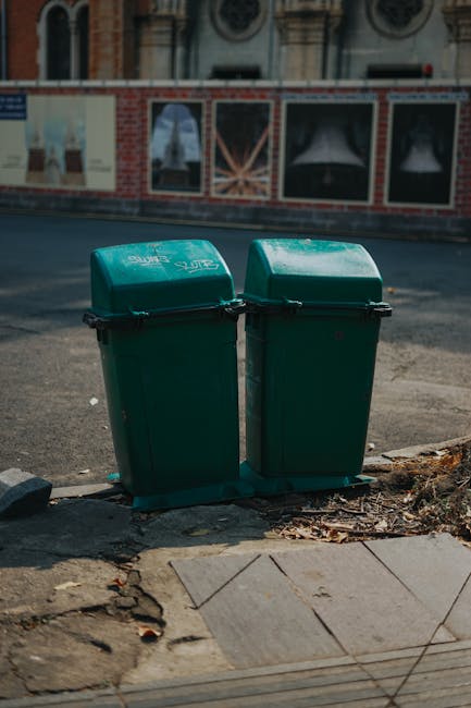 A weathered metal rubbish bin with a graffiti-styled face painted on the front, featuring dark eye sockets, a nose, and an open mouth, situated outdoors on a grassy area. The bin has a black lid, which appears slightly bent or damaged, and is partially covered with clearing bags made of plastic or fabric in shades of blue and green, placed on top. Surrounding the bin are green leaves and a few small plants growing at its base, with a backdrop of blurred trees and sunlight filtering through foliage, creating a natural outdoor setting. The environment suggests an informal or public space, possibly a park or garden, where waste disposal is handled via independent collection methods, aligning with private or alternative rubbish removal services rather than municipal curbside collection. The scene emphasizes the aspect of on-site or site-specific rubbish disposal, reflecting practices of waste management companies like Waste Disposal Lambeth that provide targeted waste removal services outside traditional council systems.